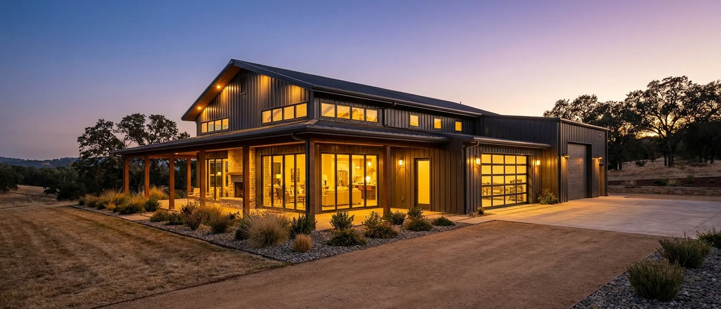A barndominium at dusk with warm light glowing from every window and exterior lighting along the porch and roofline, golden oak-dotted hills fading into the twilight behind.