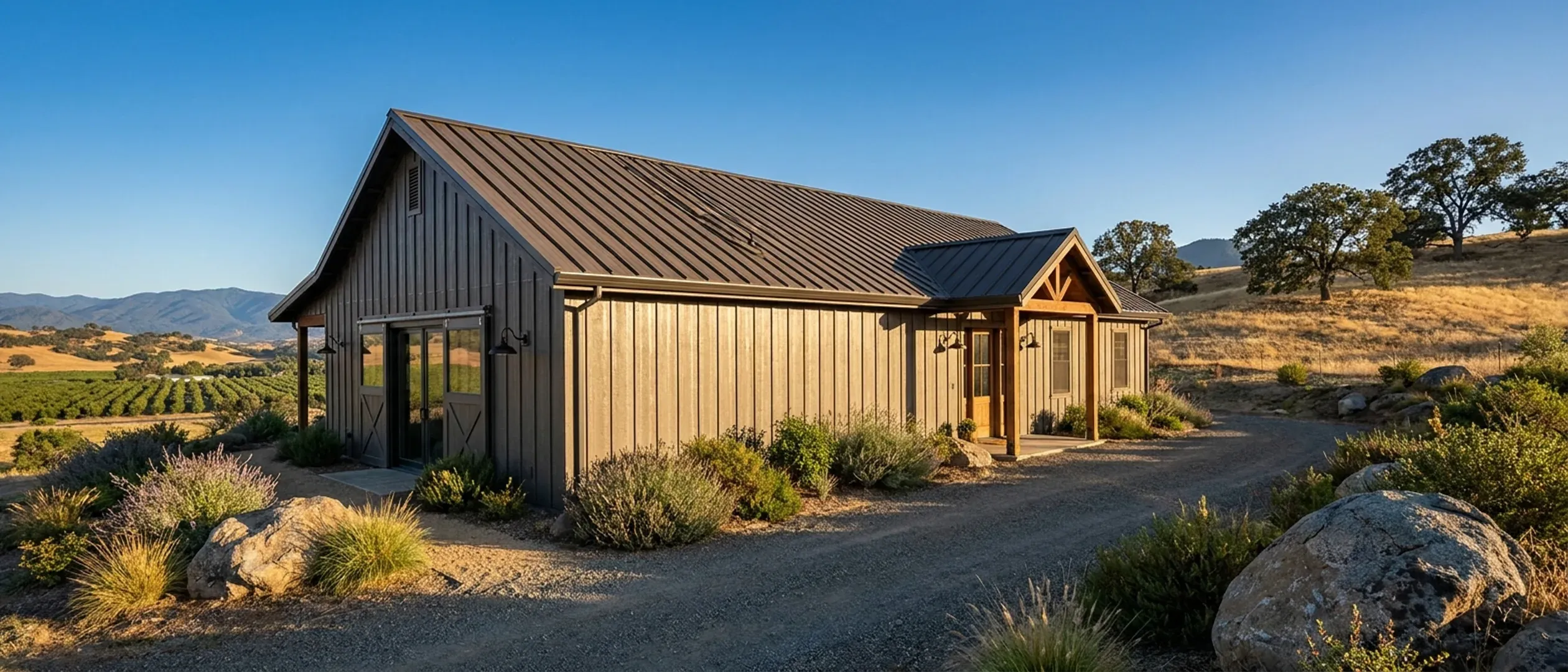 A completed metal-clad barndominium from a low angle, showing its standing seam roof, gutters and exterior wall lights against golden hills.