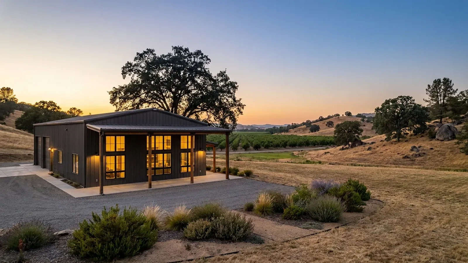 A finished barndominium at dusk with warm interior lighting glowing through its large windows and an oak silhouetted behind.