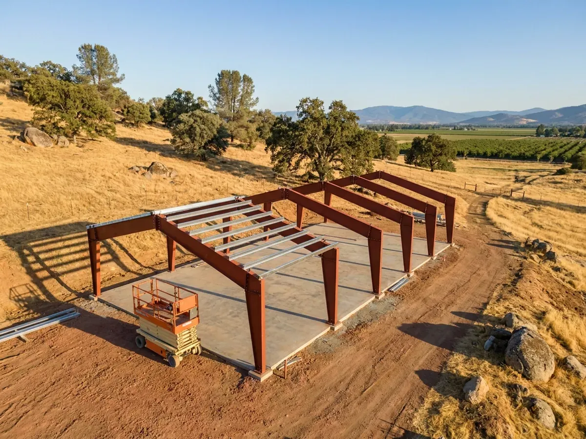 Rust-red primed steel frames standing in a row on a concrete slab, with purlins going on across the first bays and a scissor lift beside them.